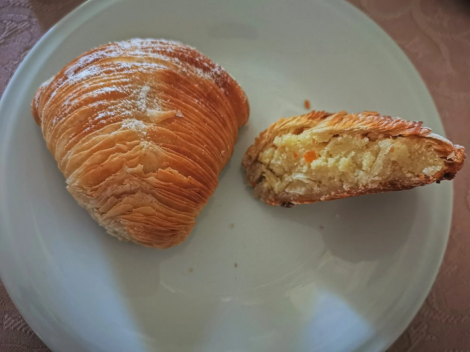 Una sfogliatella riccia spolverata di zucchero a velo su un piatto bianco, accanto a un pezzo tagliato che mostra il ripieno di ricotta e semola con canditi