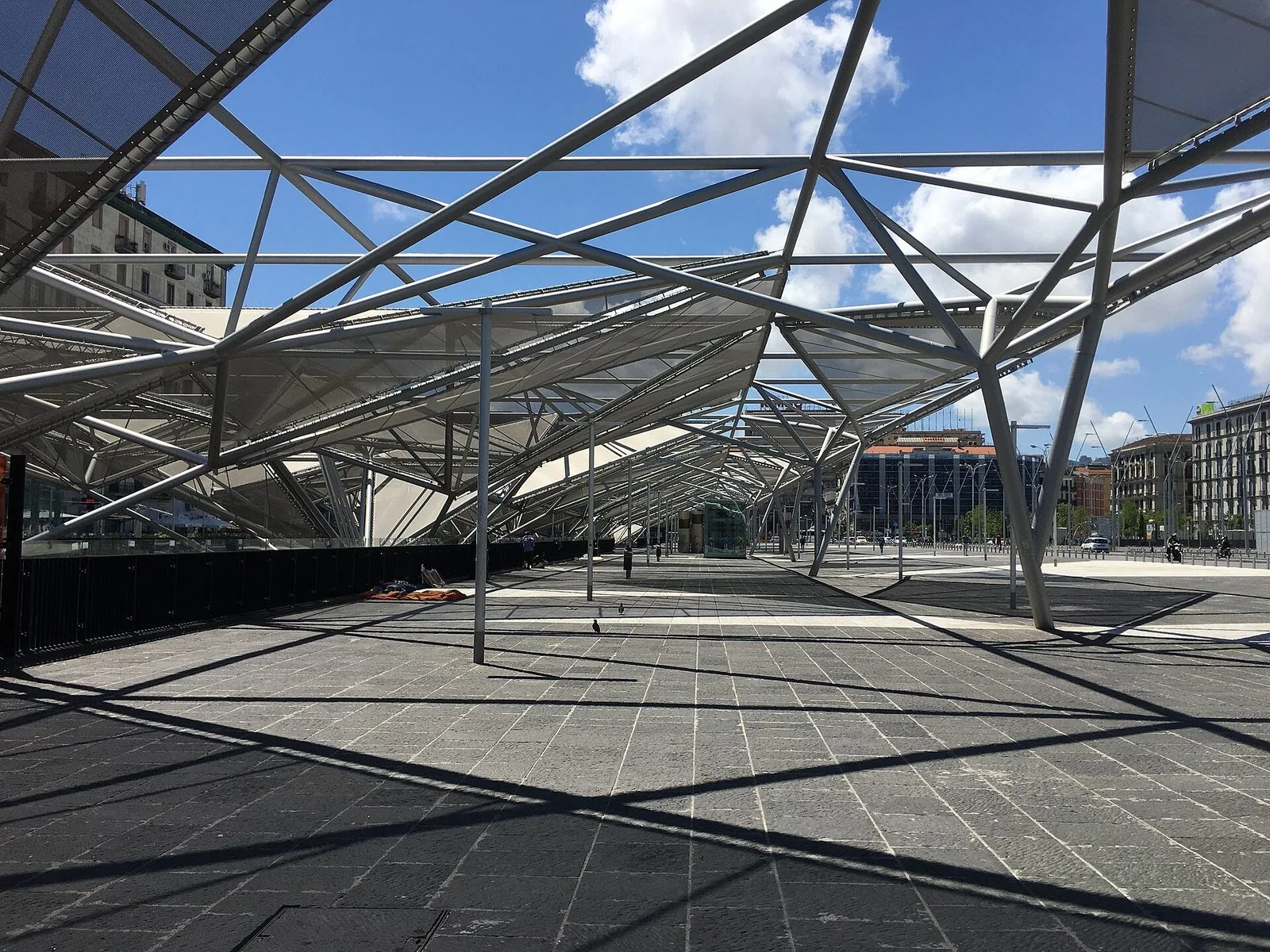 Le pensiline in acciaio e tessuto di Piazza Garibaldi a Napoli viste da sotto, con le ombre geometriche sulla pavimentazione in pietra scura, il cielo azzurro con nuvole e i palazzi della piazza sullo sfondo